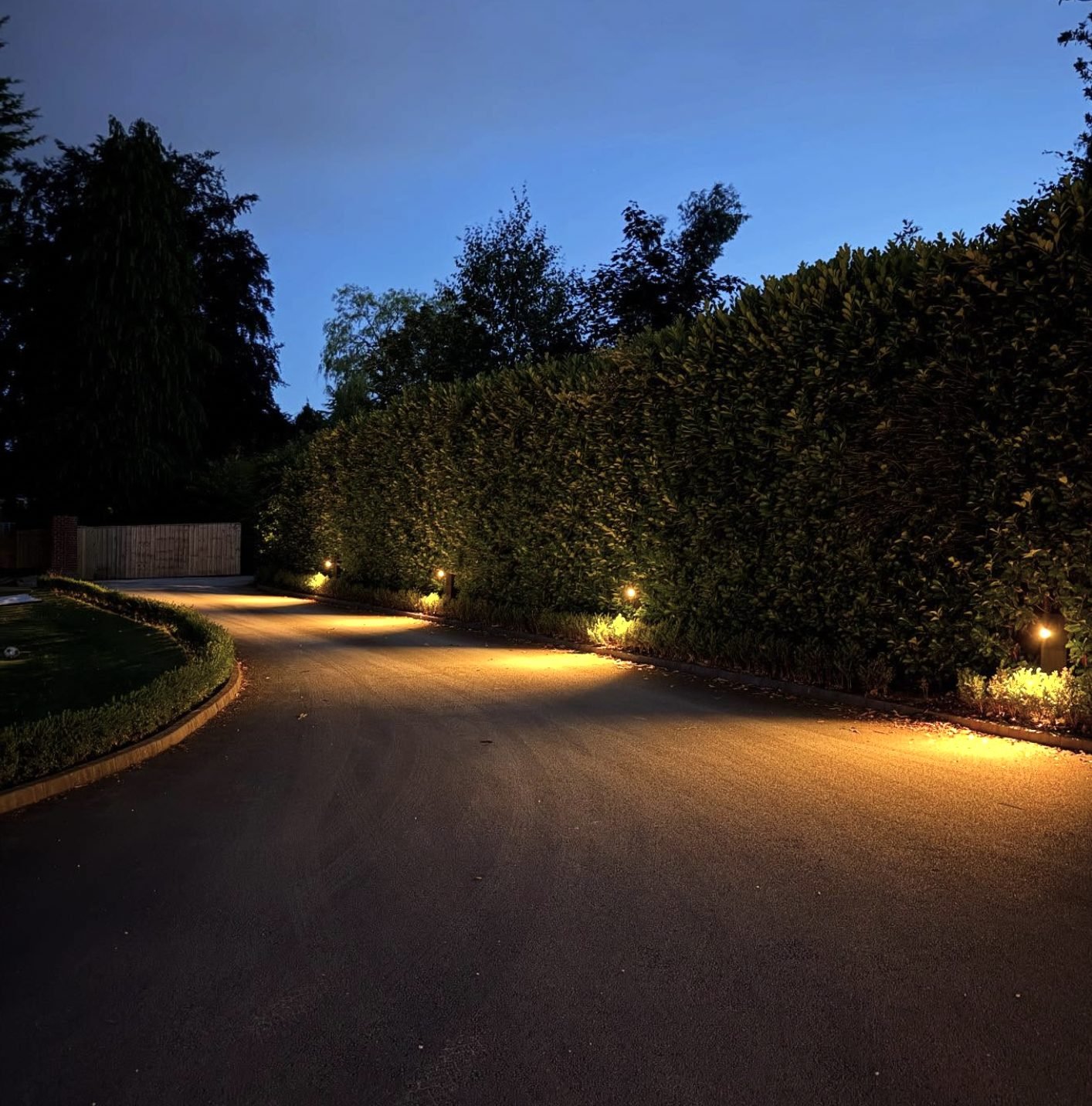 Driveway lit at dusk with warm bollard and border lighting along a hedge