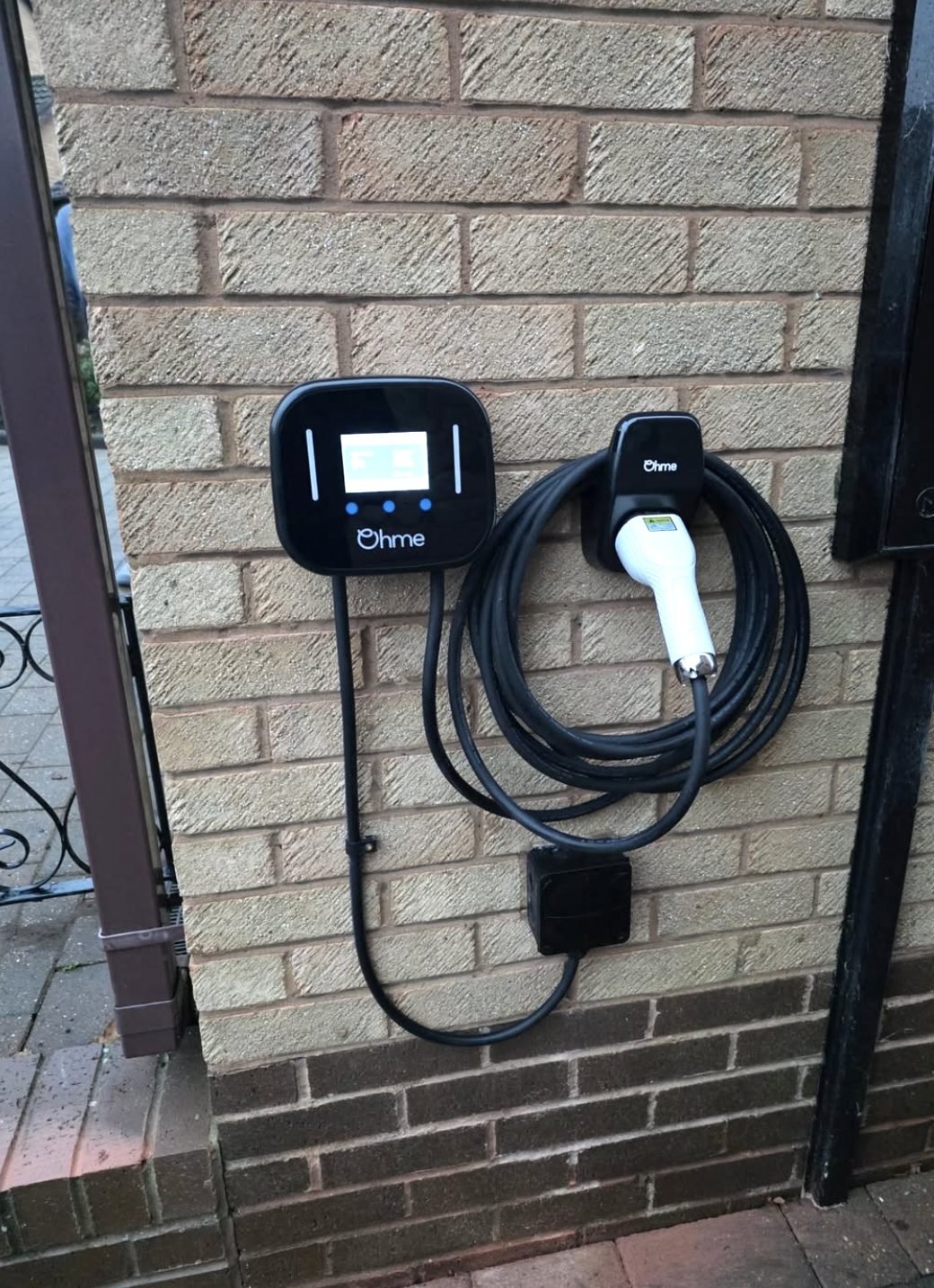 Ohme tethered EV charger with coiled cable installed beside a driveway gate